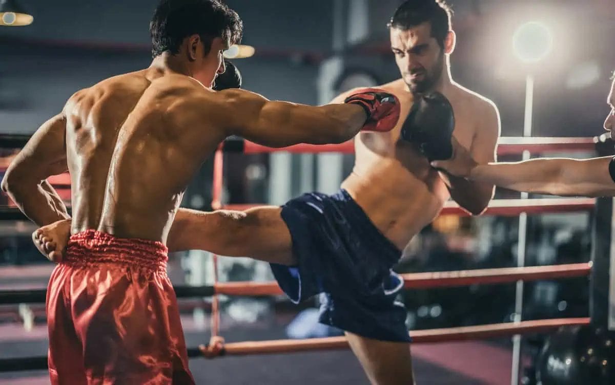 A Muay Thai boxer delivers a powerful kick to the pads held by a trainer during a practice session in a well-equipped gym
