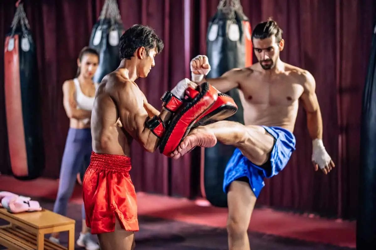A Muay Thai instructor holds pads for a male student practicing high kicks in a gym with another female student training in the background