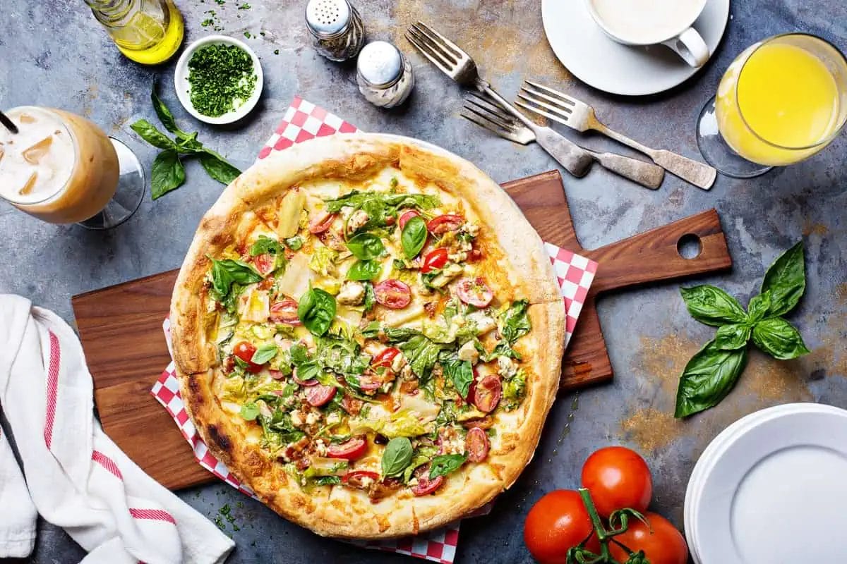 A fresh, vegetable-topped pizza on a wooden board with breakfast beverages