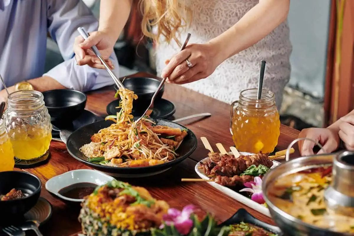 A group of people sharing a traditional Thai meal, featuring Pad Thai and skewered meats, complemented by iced drinks