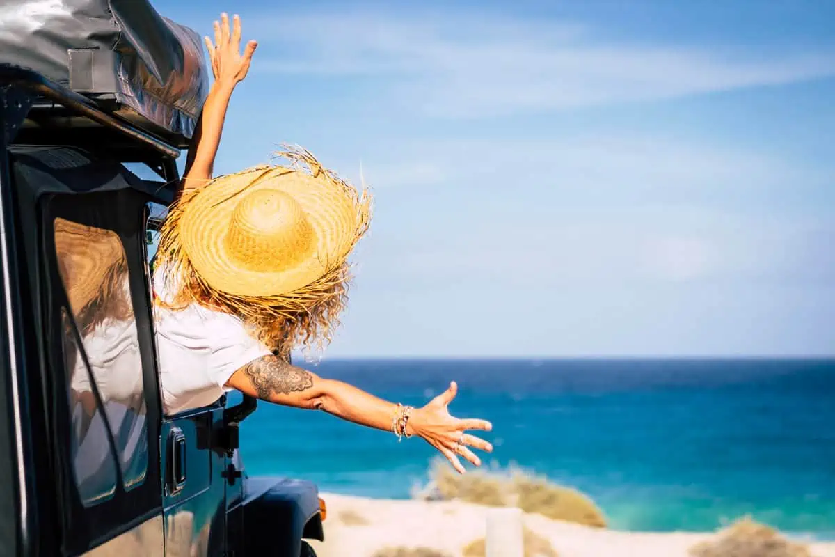 A person with a straw hat outstretched from the back of a jeep, greeting the ocean view