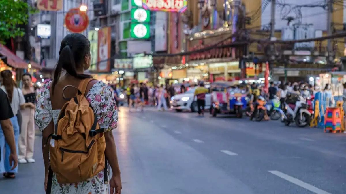 A woman with a backpack strolls through Khao San Road at evening, amidst vendors and taxis