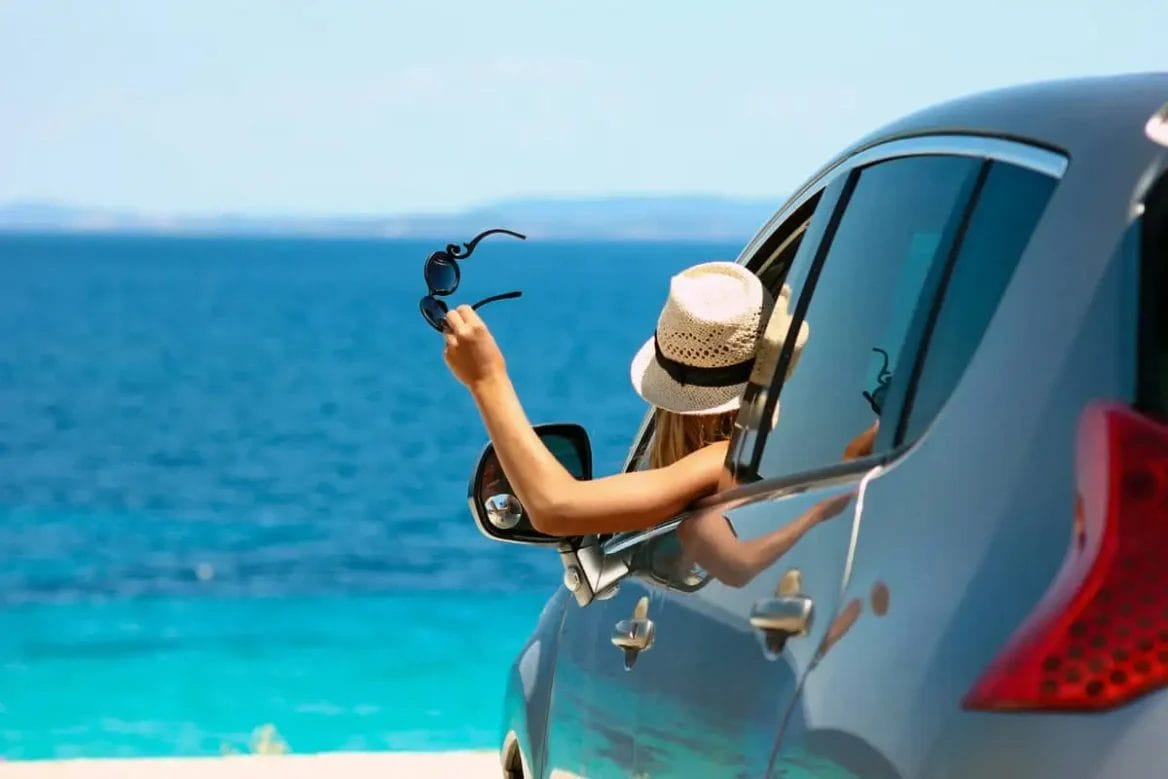 A woman's hand holding sunglasses out of a car window with a clear blue ocean in the background