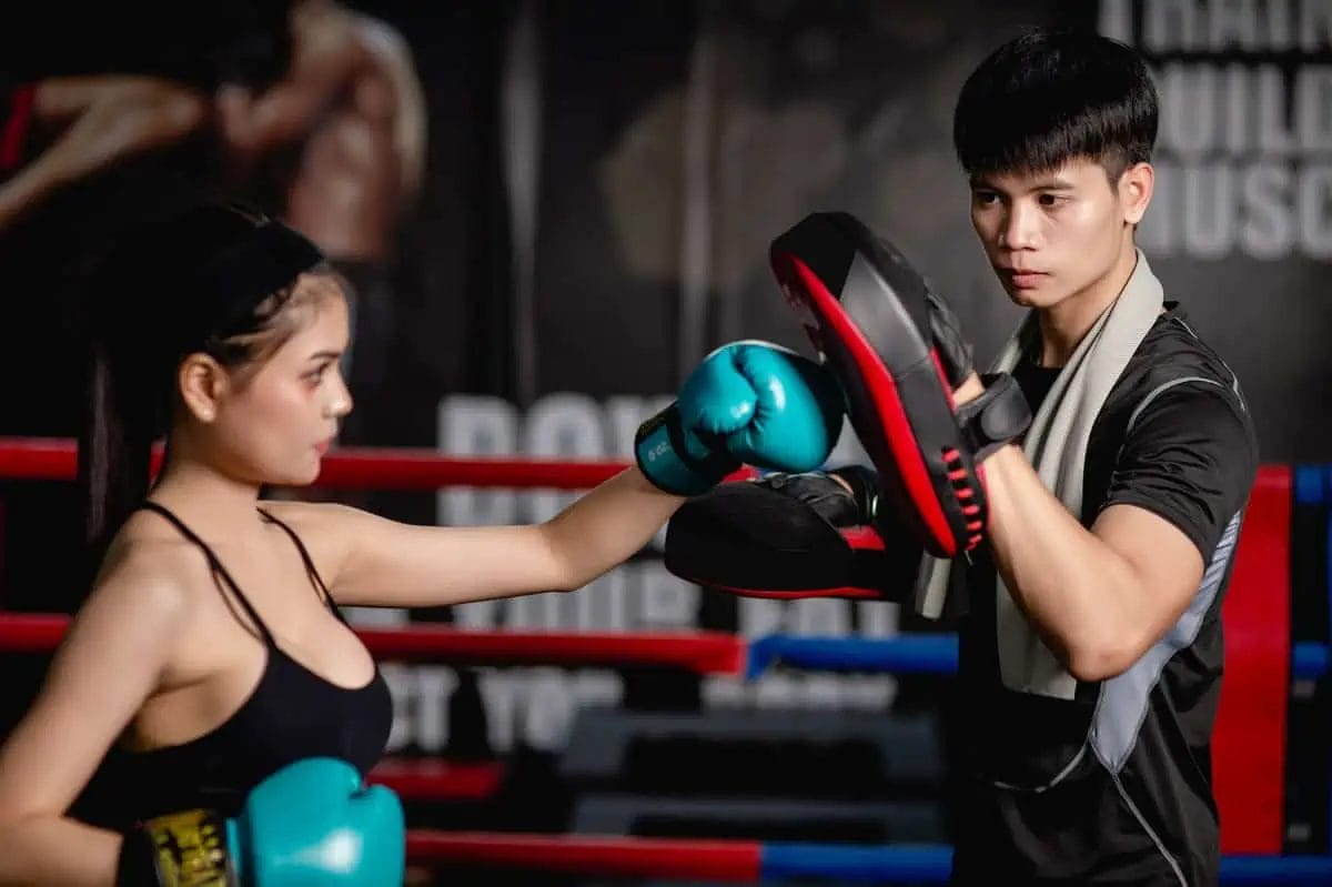 A young female boxer in blue gloves practices punching with her male coach holding focus mitts in a modern boxing gym