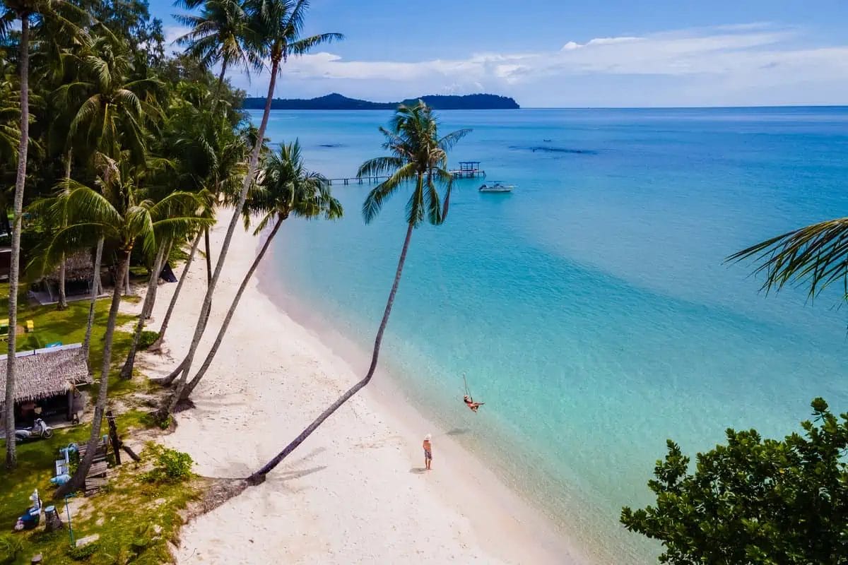Aerial view of a tranquil Thai beach with crystal clear waters, white sand, and palm trees leaning over the shore