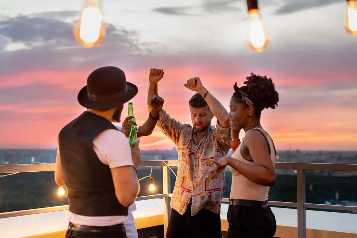 Group of friends enjoying a dance and toast with beer bottles at a rooftop bar during sunset, with city lights coming to life in the background
