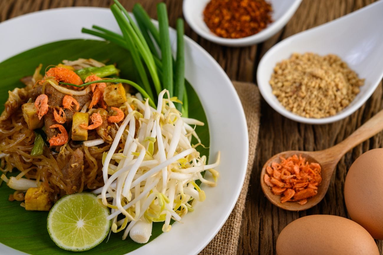 Street Food Bangkok: 5 Must-Try Dishes You Can't Miss 1 Pad Thai served on a white plate, accompanied by lemon, eggs, and seasoning, elegantly presented on a wooden table with selective focus