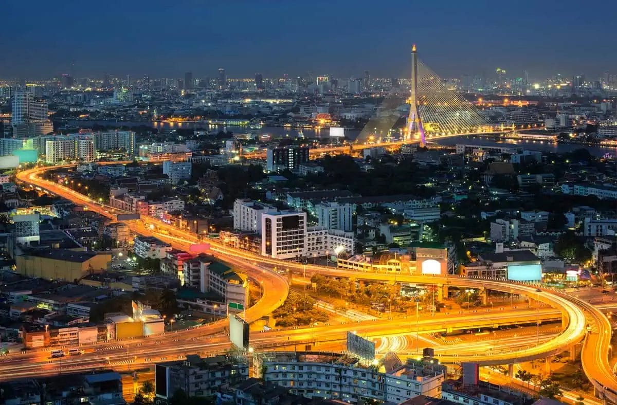 Panoramic night view of Bangkok's cityscape with a prominent bridge illuminated in purple, roads filled with light trails from moving traffic, and a dense skyline