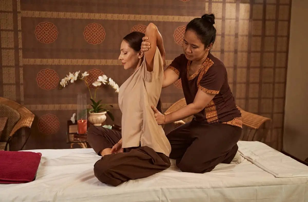Thai massage therapist stretching the arm of a seated client in a traditional spa setting
