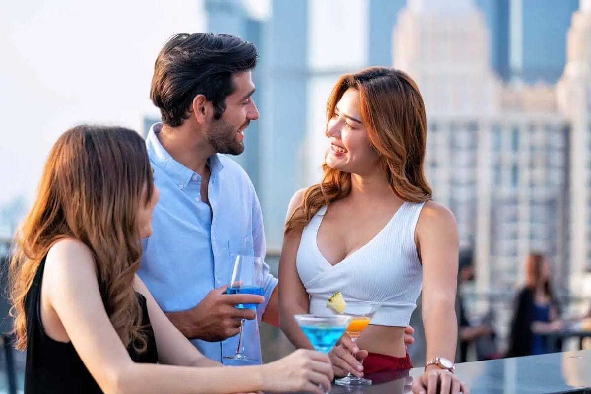 Three friends sharing a cheerful moment over blue cocktails at a rooftop bar, with the cityscape stretching behind them in soft focus
