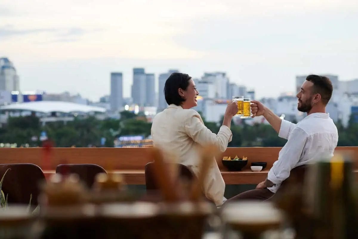 Two friends raising a toast with golden beer mugs at a rooftop venue, overlooking an expansive city view as evening sets in