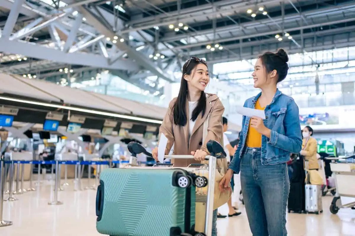 Two young women with luggage at an airport check-in area, smiling and interacting